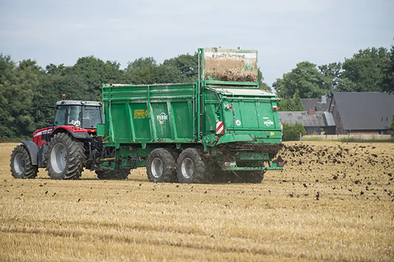 Roter Traktor mit grünem Großflächenstreuer verteilt Mist auf einem Stoppelfeld.
