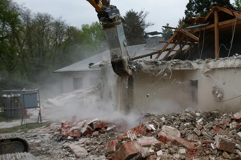 Bagger mit Abbruchgreifer reißt ein Gebäude ab, davor ein Haufen Ziegelschutt.