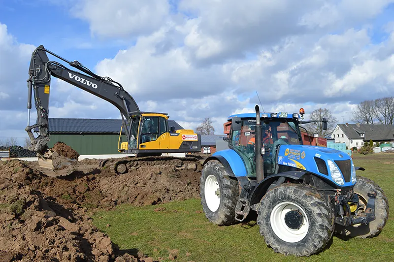 Gelber Volvo-Bagger hebt Erde aus, daneben ein blauer New-Holland-Traktor.
