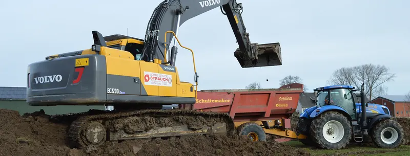Volvo-Kettenbagger belädt einen roten Kippanhänger hinter einem blauen Traktor.
