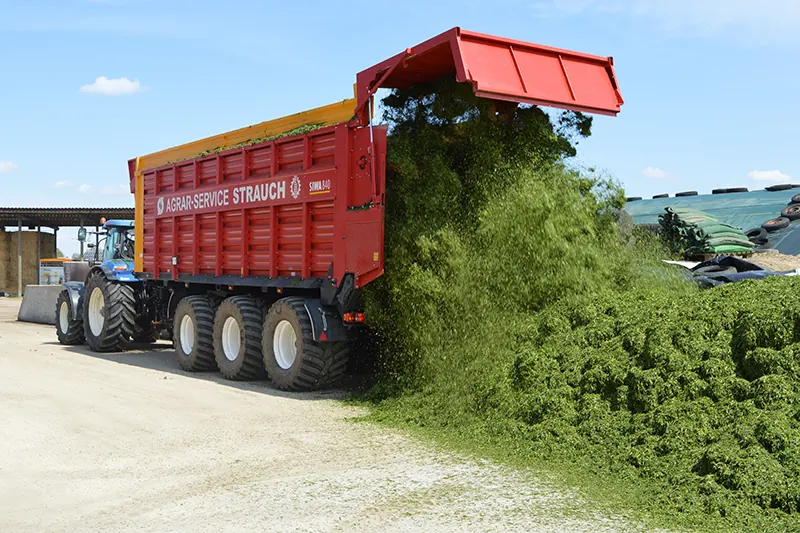 Der rote Häckselwagen von Agrar-Service Strauch kippt frisches Gras am Silo ab, davor ein blauer Traktor.