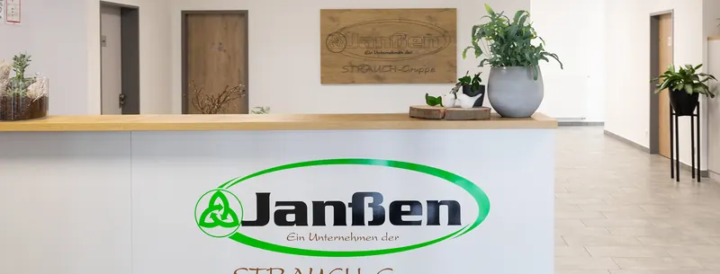 Empfangstheke mit grünem Janßen-Logo in einem hellen Flur, an der Wand ein Holzschild mit dem Logo.