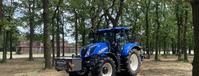 Blauer New-Holland-Traktor mit Janßen-Frontgewicht, am Heck ein Gerät mit nach oben gerichtetem Rohr, in einem lichten Wald.