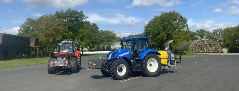 Ein roter und ein blauer Traktor mit Janßen-Frontgewichten auf einer Asphaltfläche, am blauen Traktor ein gelber Tank und ein nach oben gerichtetes Rohr.