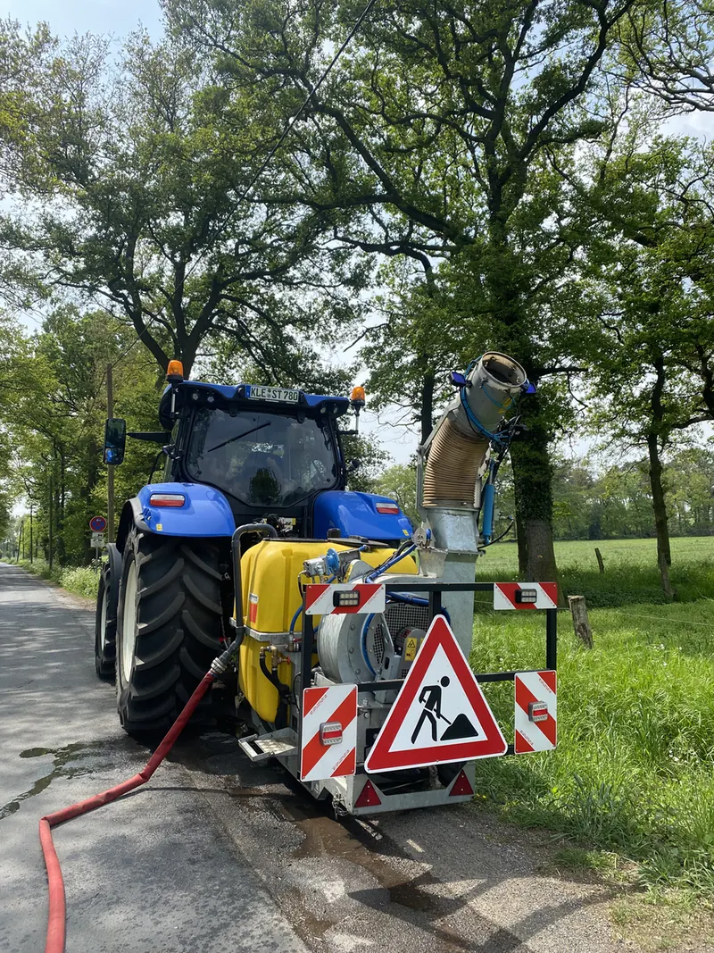 Blauer Traktor von hinten mit gelbem Tank, Gebläse mit nach oben gerichtetem Rohr und Baustellen-Warnschild am Rand einer Straße unter großen Bäumen; ein roter Schlauch liegt auf der Fahrbahn.