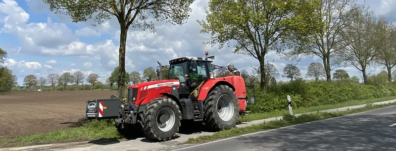 Roter Massey-Ferguson-Traktor mit Frontgewicht am Straßenrand vor einer Baumreihe, links ein Acker.