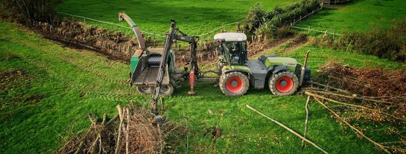 Luftaufnahme: grüner Claas-Traktor mit roten Felgen, Kran und Holzhacker zwischen Astschnitt auf einer Wiese.