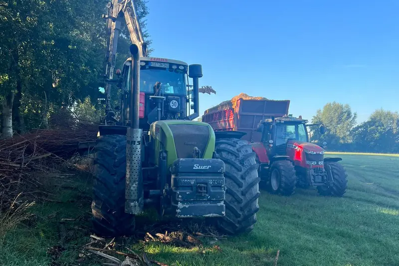 Grüner Claas-Traktor mit Kran von vorn neben einem Haufen Astschnitt, dahinter ein roter Traktor mit einem Anhänger voller Hackschnitzel.