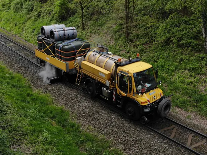 Gelber Zweiwege-Unimog mit Tank und angehängtem Wagen voller Behälter auf einem Gleis, unter dem Wagen steigt Dampf auf; Blick von schräg oben.