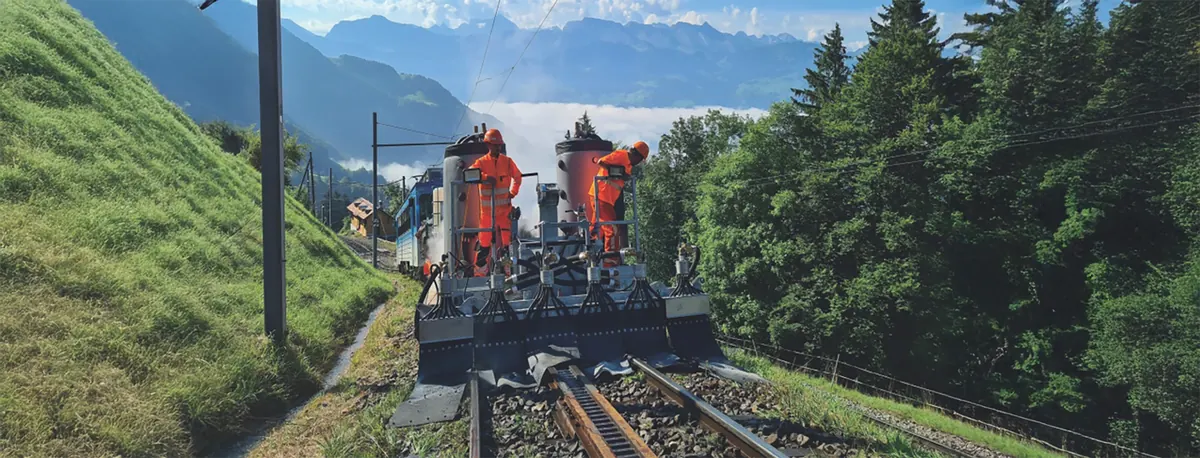 Schienenfahrzeug mit Tanks und vorn angebautem Sprühbalken auf einem Gleis mit Zahnstange im Gebirge, darauf zwei Arbeiter in orangefarbener Schutzkleidung; im Hintergrund Berge und Nebel im Tal.