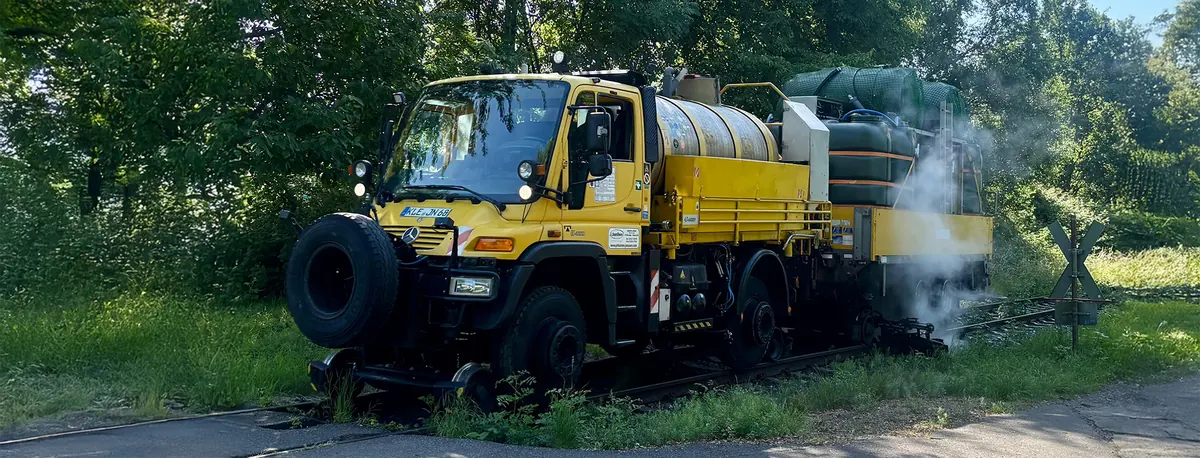 Gelber Zweiwege-Unimog mit Tank auf einem Gleis, am Heck steigt Dampf auf.