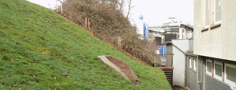 Steile, mit Gras bewachsene Böschung neben einem Gebäude, am oberen Rand eine Treppe mit Geländer; im Hintergrund Industrieanlagen.