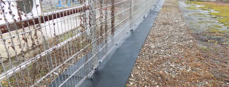 Dunkle Matte unter einem Stabgitterzaun, daneben Kies.