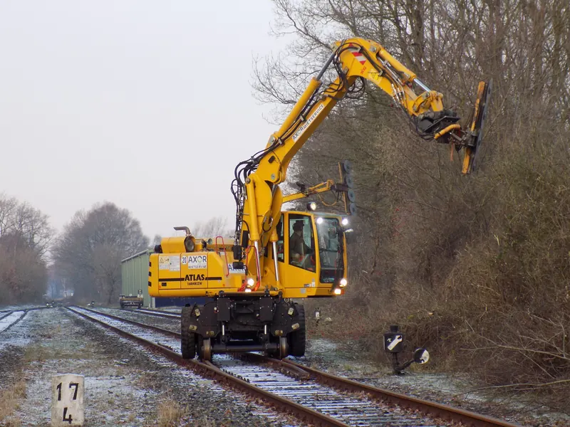 Gelber Zweiwegebagger auf dem Gleis schneidet im Winter mit einem Mulchgerät am Ausleger Gehölz am Streckenrand, Raureif liegt auf dem Schotter.