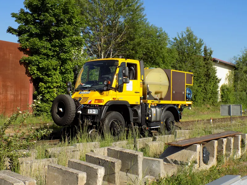 Gelber Zweiwege-Unimog mit Tank auf einem Gleis hinter einer Reihe Betonblöcke, dahinter Bäume.