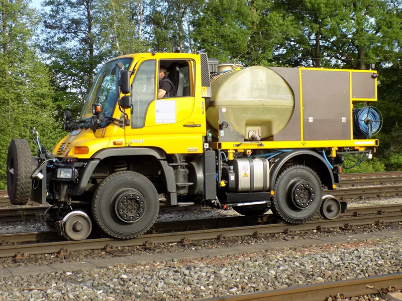 Gelber Zweiwege-Unimog mit Tank und Schlauchtrommel auf Schienenrädern, in der Kabine ein Fahrer.