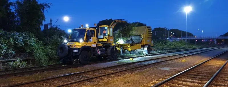 Gelber Zweiwege-Unimog mit Kran und angehängtem Schredder in der Abenddämmerung auf dem Gleis, beleuchtet von Scheinwerfern und Straßenlaternen.