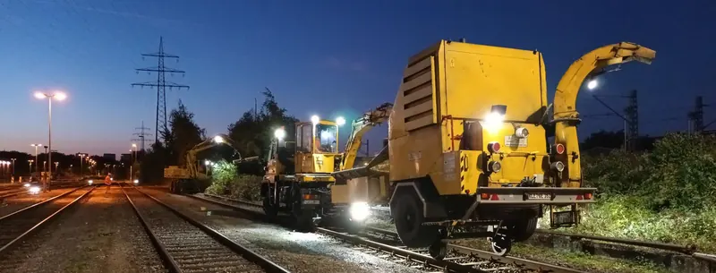 Zweiwegebagger und gelber Schredder mit Auswurfrohr auf dem Gleis in der Dämmerung, von Scheinwerfern beleuchtet, im Hintergrund Strommasten.