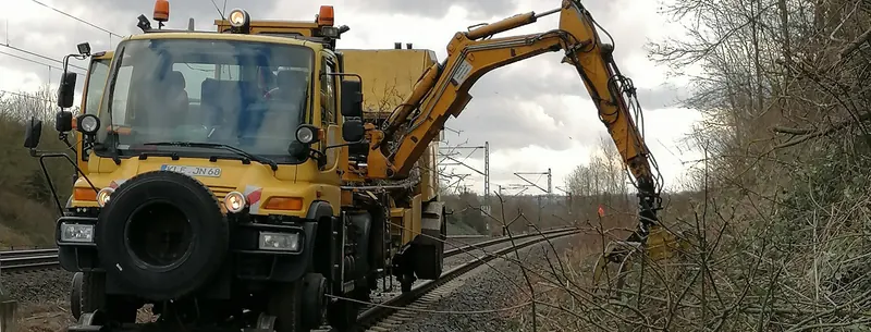 Gelber Zweiwege-Unimog von vorn, mit dem orangefarbenen Auslegerarm schneidet er Sträucher neben dem Gleis.
