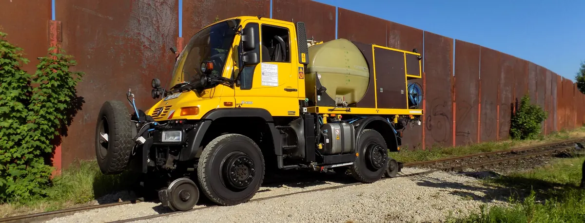Gelber Zweiwege-Unimog mit Tank und Schlauchtrommel auf einem Gleis vor einer rostbraunen Wand.