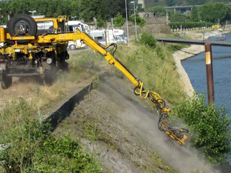 Gelbes Fahrzeug mit einer Bürste am langen Auslegerarm bearbeitet eine schräge Ufermauer an einem Fluss, dabei wirbelt Staub auf.