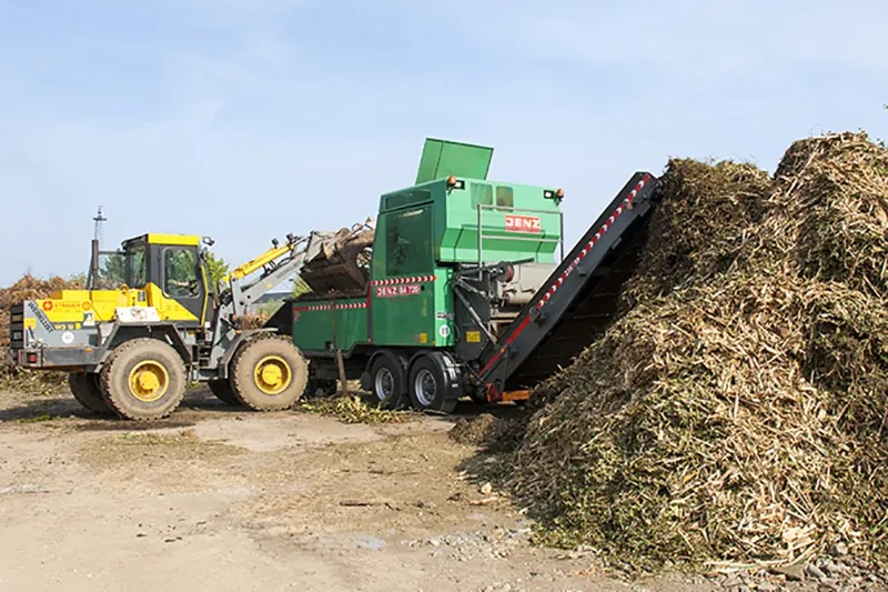 Grüner Jenz-Biomasseaufbereiter, beschickt von einem gelben Radlader, daneben Haufen mit Häckselgut.