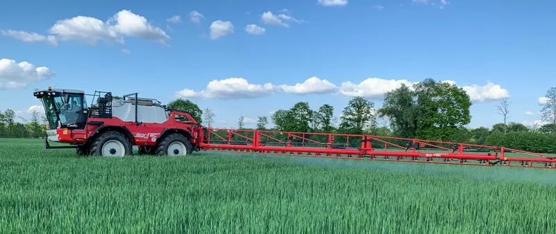 Rote selbstfahrende Pflanzenschutzspritze mit weit ausgeklapptem Gestänge über einem grünen Getreidefeld.